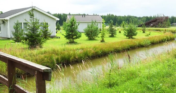 Wooden bridge railing over narrow water canal near modern countryside cottages. Scenic residential estate featuring young pine trees on green grassy lawn. Beautiful eco resort village with white woode