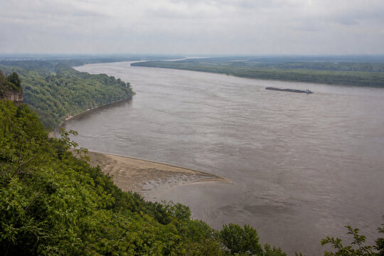 View from high bluff of wide brown river bending through low green forest, sandbar, tugboat and barge downstream, Mississippi River landscape