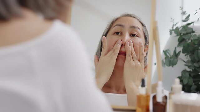 Asian mature woman with natural grey hair applies moisturizing facial serum to her cheek while looking in a mirror. Slow-motion shot.