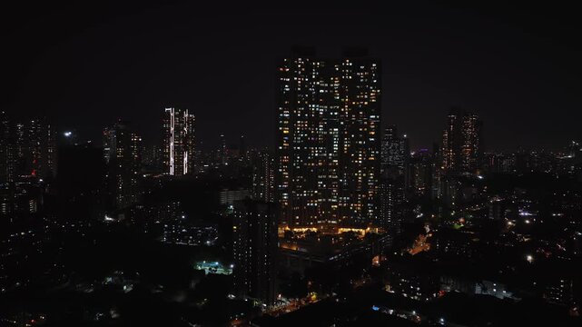 An aerial view of Mumbai's high-rise buildings and beautiful nightlights. The sky is cloudy, and there is medium traffic on the road. The cityscape is filled with modern illuminated buildings