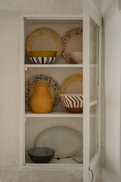 Open White Cabinet Displaying Handcrafted Ceramic Plates Bowls And Yellow Pitcher In Rustic Mediterranean Kitchen
