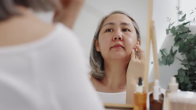 Asian mature woman with natural grey hair applies moisturizing facial serum to her cheek while looking in a mirror. Slow-motion shot.