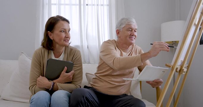 Senior husband paints nature artwork on canvas while senior wife holds tablet reference and watches nearby at home, combining creative therapy, companionship, relaxation, and active retirement.