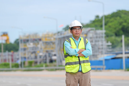 Engineer, construction, hard hat, safety vest, industrial site, worker standing with crossed arm near road, confident Asian man at outdoor project area under blue sky