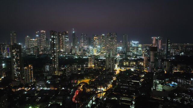 An aerial view of Mumbai's high-rise buildings and beautiful nightlights. The sky is cloudy, and there is medium traffic on the road. The cityscape is filled with modern illuminated buildings