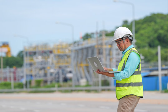Engineer worker helmet safety vest laptop industrial site inspection, focused male professional using computer near refinery structure during outdoor maintenance work
