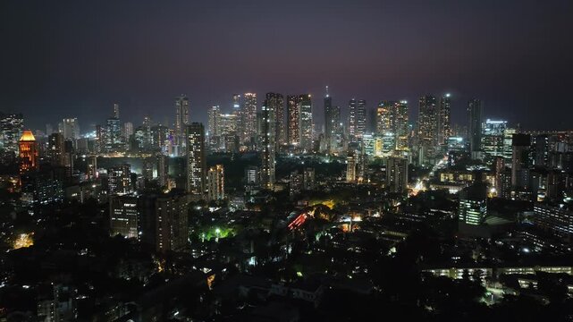 An aerial view of Mumbai's high-rise buildings and beautiful nightlights. The sky is cloudy, and there is medium traffic on the road. The cityscape is filled with modern illuminated buildings