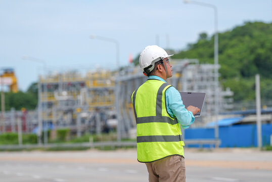 Engineer worker hardhat safety vest tablet, industrial plant inspection outdoors, male technician checking infrastructure near road with focused posture during daytime