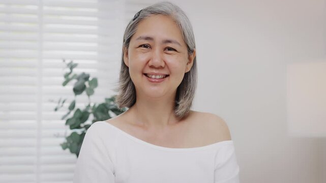 Slow-motion Asian mature woman with natural grey hair looks at the camera with a gentle, confident smile.