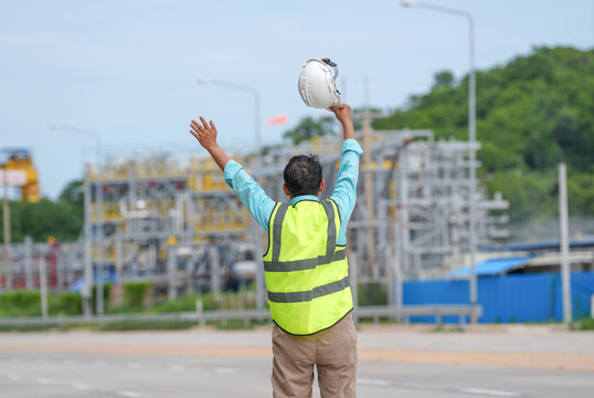 Construction worker safety vest hard hat industrial site raised arm road back view construction worker wearing safety vest lifting hard hat with raised arm near road and industrial site