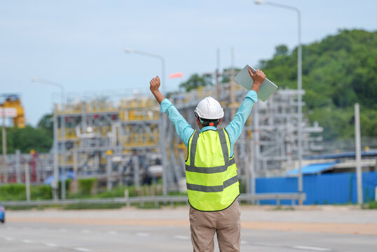 Engineer worker hard hat safety vest tablet construction site industrial plant road success gesture behind male supervisor facing refinery project area outdoors