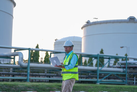 Engineer helmet safety vest laptop oil tank pipeline industrial site male worker inspection at storage facility with focused expression near metal pipe during daylight
