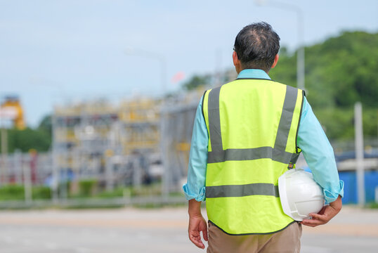 Engineer safety vest hard hat industrial site construction worker male outdoor back view inspection near road with equipment green tree calm professional stance