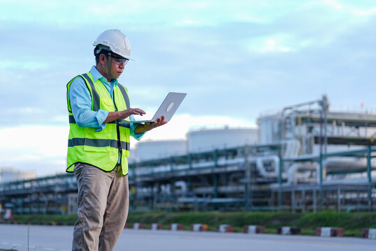 Engineer, hard hat, safety vest, laptop, refinery, industrial plant, worker using computer near pipeline equipment during outdoor inspection under cloudy sky