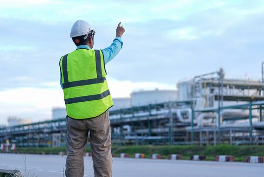 Engineer hardhat safety vest industrial plant pipeline worker pointing toward refinery infrastructure during outdoor inspection under cloudy sky with focused posture