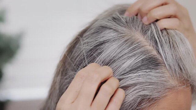An extreme close-up of a mature Asian woman parting her hair, showcasing natural grey roots and authentic hair texture.