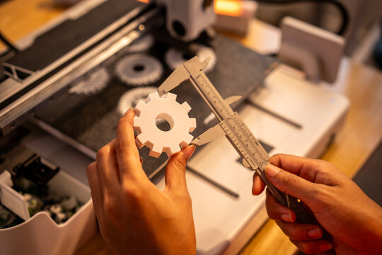 Hands measuring a white 3d printed gear part with a caliper near a desktop printer in a workshop