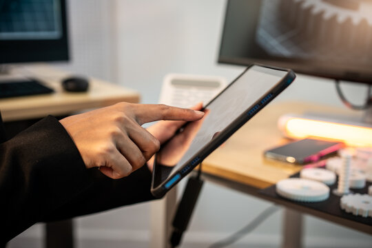 Hand using a tablet on a desk in a modern office with computer screens and warm light, showing a simple work scene