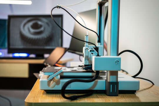 Desktop 3d printer on a wooden table in a workshop or office, with cables and a computer monitor in the background