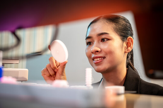 Smiling young woman examining a small round sample at a desk in a modern office, with soft colorful lights in the background