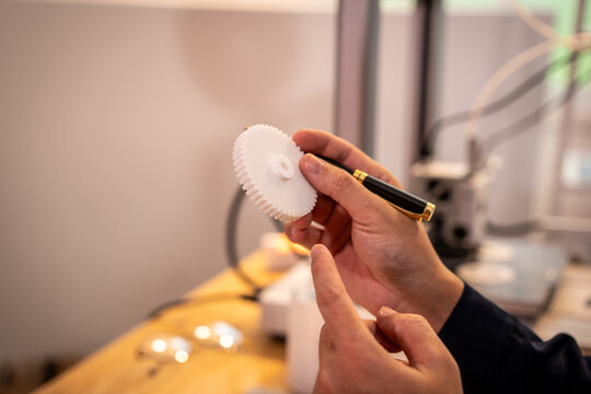 Close-up of a hand holding a white plastic gear and pointing at it with a pen in a workshop, showing design inspection and technical work.