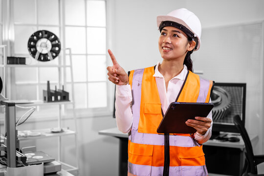 Young female engineer in a hard hat and safety vest holding a tablet and pointing in a modern office, working on technical plans