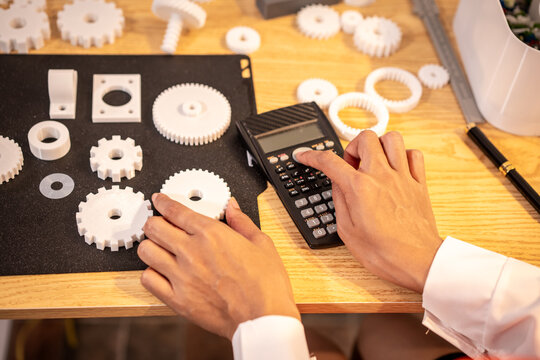 Hands using a calculator beside plastic gears and parts on a wooden desk, showing engineering planning, design work and problem solving.