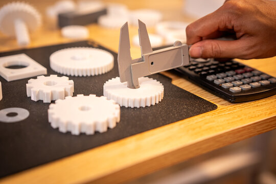 Hand measuring a white plastic gear with caliper beside calculator and other 3d printed parts on a workbench.