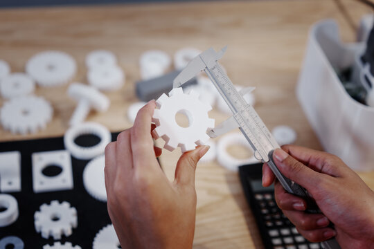 Hands measuring a white plastic gear with calipers on a workbench, with other parts and tools blurred in the background