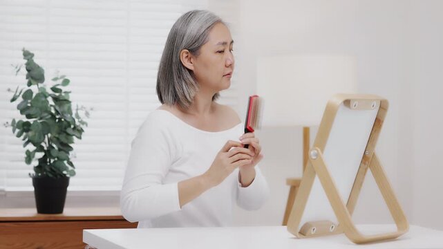 An Asian mature woman with natural grey hair gently brushes her short hair while looking into a tabletop mirror, Slow-motion shot.