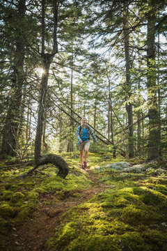 Smiling woman hiking on a sunlit mossy forest trail in Maine.