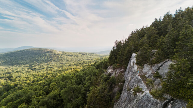 Solo hiker on a cliff overlooking a vast forest in Maine woods.