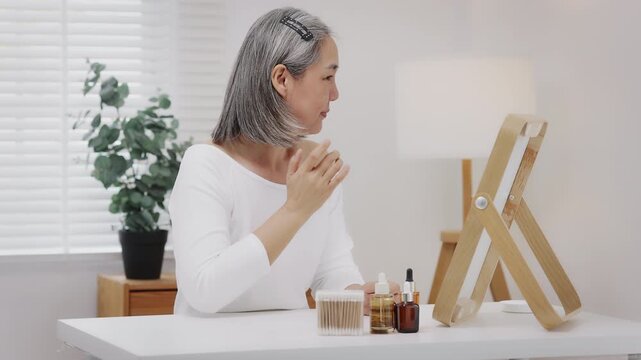 Asian mature woman with natural grey hair applies a moisturizing facial serum to her cheek while looking in a mirror. Slow-motion shot.