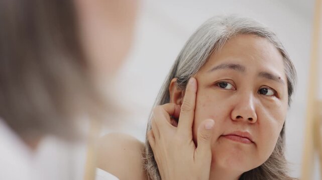 An Asian mature woman with grey hair looks in a mirror with concern while checking fine lines and aging skin.