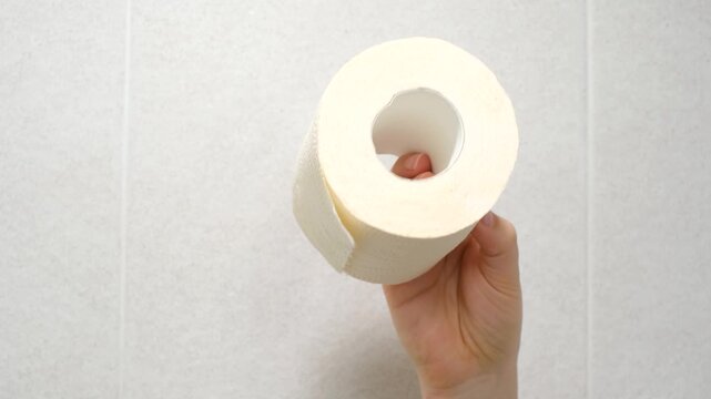 Hand holding a roll of toilet paper on gray tile background close-up.