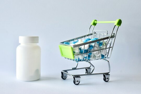 Miniature shopping cart filled with medical capsules and tablets placed next to a blank white medicine bottle, representing healthcare costs, pharmaceutical shopping, and medication expenses.
