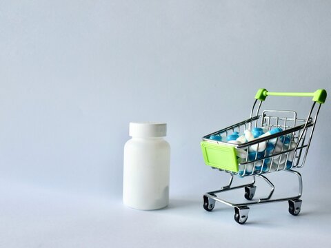 Miniature shopping cart filled with medical capsules and tablets placed next to a blank white medicine bottle, representing healthcare costs, pharmaceutical shopping, and medication expenses.