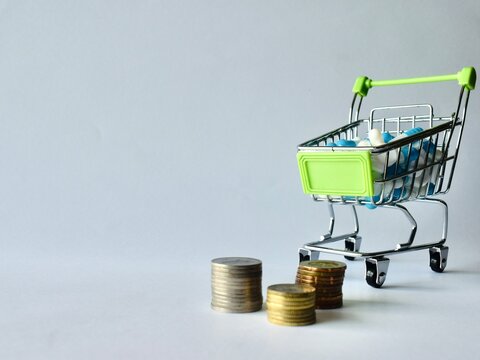 Mini shopping trolley filled with blue and white capsules positioned next to three stacks of coins representing pharmaceutical costs.