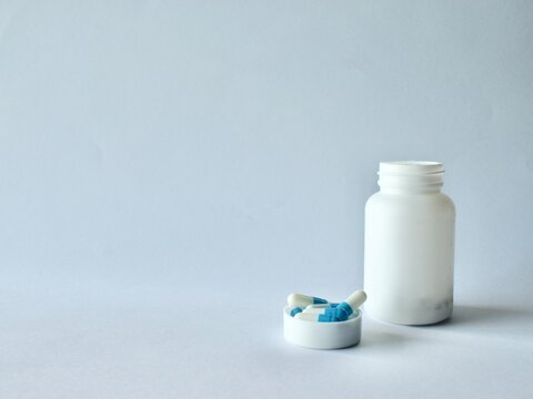 White plastic pill bottle placed next to a container cap filled with blue and white medicinal capsules on a clean neutral backdrop.