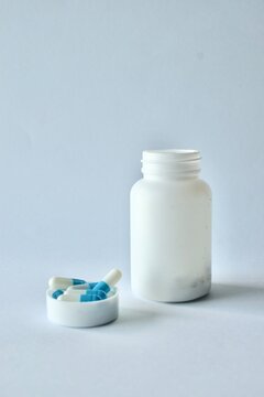 White plastic pill bottle placed next to a container cap filled with blue and white medicinal capsules on a clean neutral backdrop.