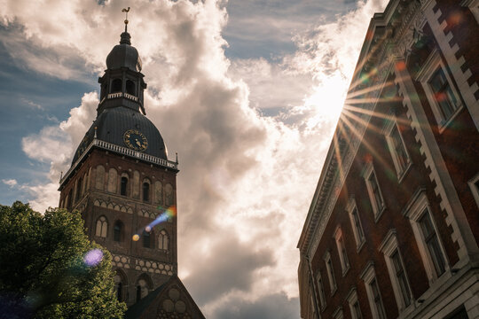 Low-angle view of the historic Riga Cathedral bell tower with a bright sunburst flaring through clouds over old town street in Riga, Latvia