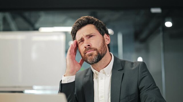 Professional experiences intense headache at modern office workstation. Symptoms of fatigue and frustration due to work stress and overload are evident. Discomfort impacts concentration productivity.