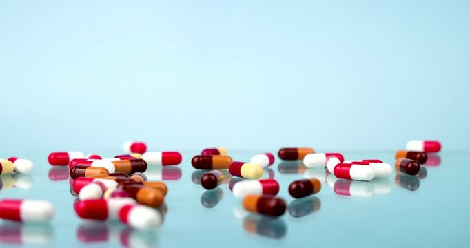 Medical capsules in red, white, orange, and brown dropping onto a clean light blue glass table, prescription medication, pharmacy treatment, and health care concept display