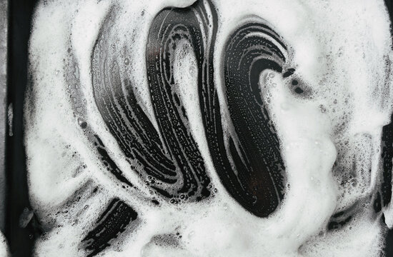 White foam and soap on dark background. The concept of washing oily pan or black car. Abstract pattern.