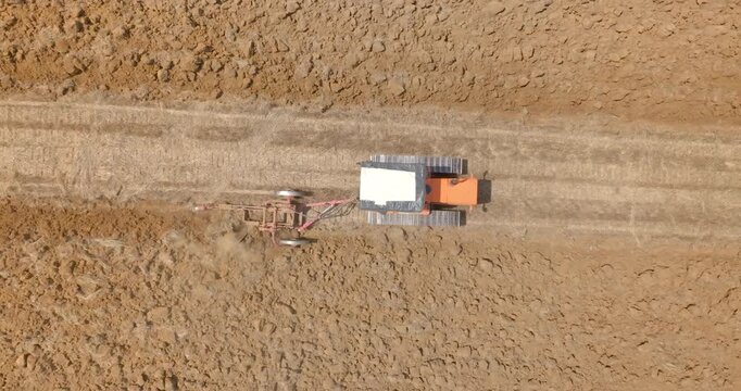 Aerial zenithal closeup of a orange tractor plows a agricultural field. The bulldozer moves across a dry dirt field, towing a long metal agricultural attachment behind it. Perpendicular view.
