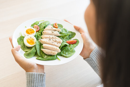 Close up of a woman holding and presenting a white plate of healthy fitness salad topped with sliced chicken breast, boiled egg and fresh tomatoes on her hand at home, weight loss meal concept.