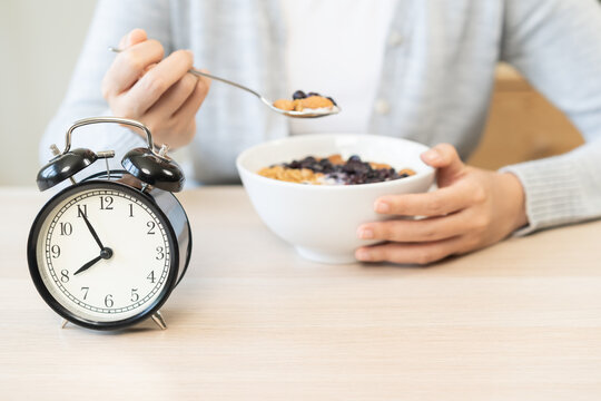 Young woman eating a healthy Greek yogurt bowl with granola and blueberries at a wooden table in the morning, with alarm clock showing the breakfast time.