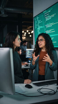 Two women in office discuss code on large screen, symbolizing teamwork, tech, and innovation