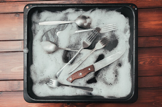 A wooden knife, fork and spoons in the detergent foam on a black oven-tray. Washing dishes concept.