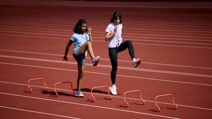 Fototapeta premium Teenage female runner practicing high knee hurdle drill beside adult female coach. Concept of agility and speed training, junior hurdle practice, summer sport coaching.